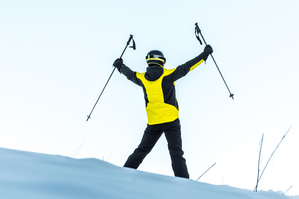achteraanzicht van de skiër in helm holding sticks terwijl hij op witte sneeuw staat  - Foto, afbeelding