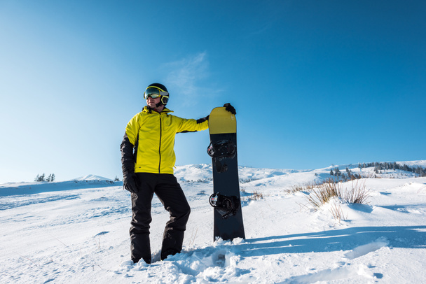 sportman in helm met snowboard terwijl hij op witte sneeuw staat  - Foto, afbeelding