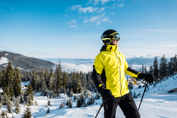 szczęśliwy narciarz w goglach trzymający kije narciarskie przeciwko błękitnemu niebu w górach  - Zdjęcie, obraz