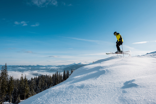 side view of skier holding ski sticks and jumping against blue sky in mountains  - Fotografie, Obrázek