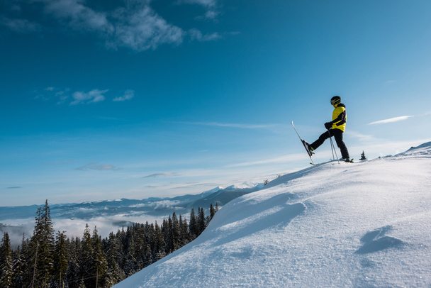 πλαϊνή όψη σκιέρ που κρατά μπαστούνια του σκι και κάνει βήμα κατά του γαλάζιου ουρανού στα βουνά  - Φωτογραφία, εικόνα