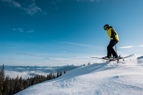 боковой вид лыжника, держащего лыжные палки во время прыжка на голубом небе в горах
  - Фото, изображение