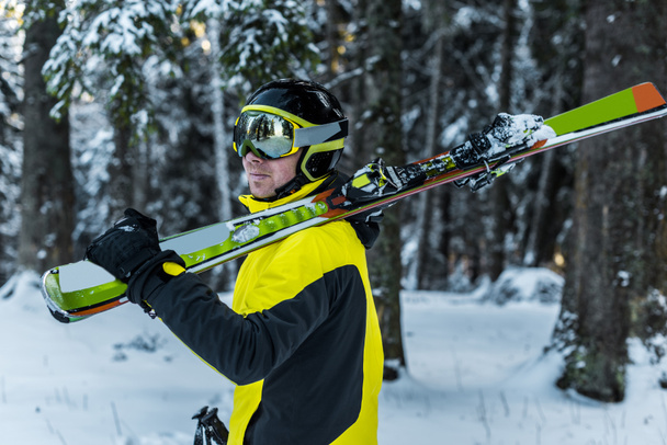 Skifahrer mit Skibrille hält Skistöcke und Skier in der Nähe von Tannen  - Foto, Bild