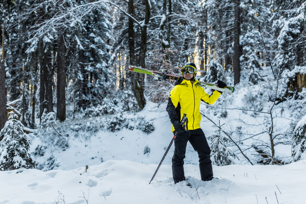 narciarz w goglach trzymający kije narciarskie i narty i stojący w pobliżu firs  - Zdjęcie, obraz
