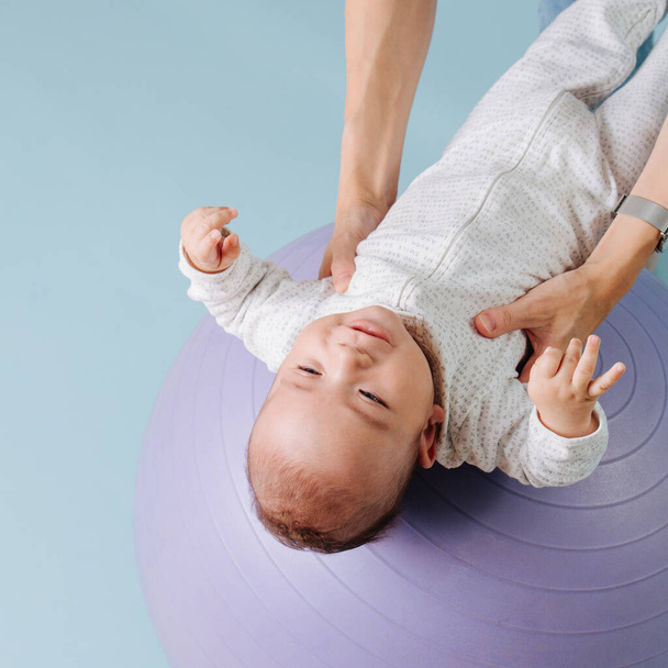 Малыш в белом комбинезоне лежит на сиреневом фитнес-мяче
 - Фото, изображение