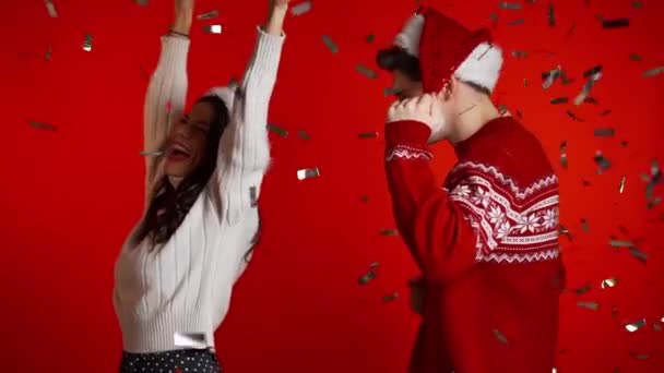 Excited couple Santa Claus hats dancing, applauding, having fun, rejoices over confetti rain in red studio. Concept of Christmas, New Year, happiness, party, winning. - Footage, Video