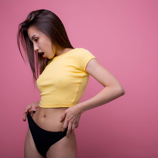 Beautiful Asian woman posing and showing how thin she pulls black panties in f yellow t-shirt on a pink background - Photo, image