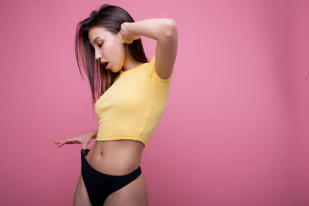 Beautiful Asian woman posing and showing how thin she pulls black panties in f yellow t-shirt on a pink background - Photo, image