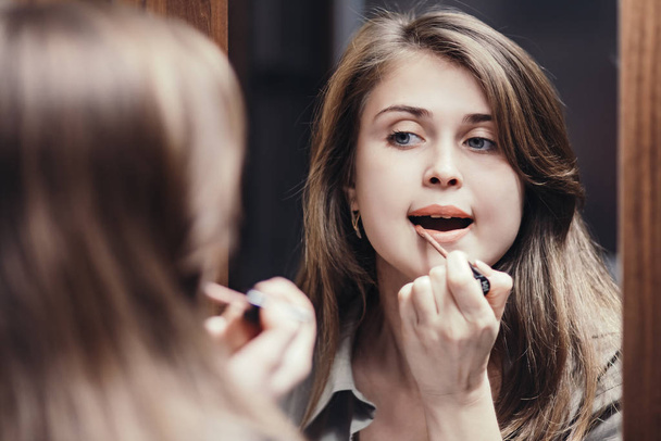 girl paints her lips with lipstick in front of a mirror at home, a young woman doing makeup, a concept of female natural beauty and cosmetics - 写真・画像