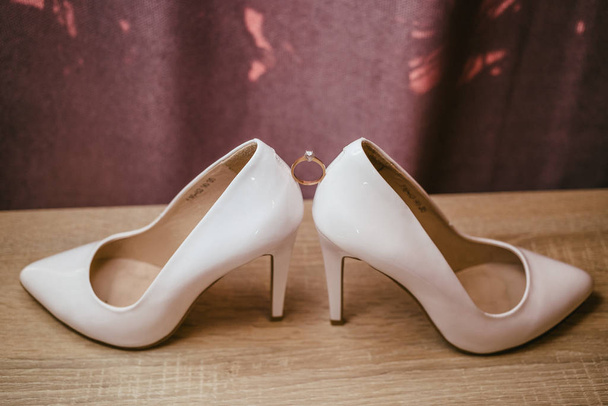 Χρυσό διαμαντένιο δαχτυλίδι μεταξύ δύο λευκών ψηλοτάκουνων παπουτσιών. Λεπτομέρειες γάμου σε κόκκινο φόντο - Φωτογραφία, εικόνα