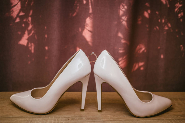 Gold diamond ring between pair of white high heel shoes. Wedding details on red background - Photo, Image