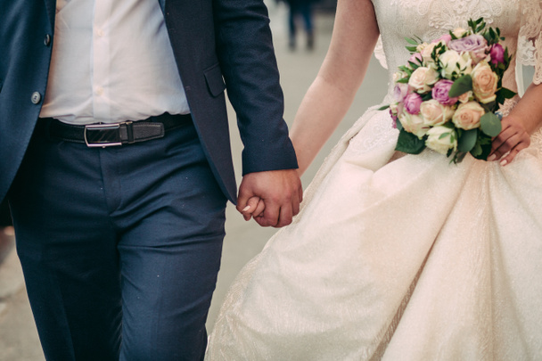 Γαμήλιο μπουκέτο στα χέρια του γαμπρού και της νύφης. Το ζευγάρι περπατά κρατώντας τα χέρια. Με λευκό φόρεμα και μπλε σακάκι - Φωτογραφία, εικόνα