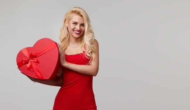 A young woman in a red dress blows a kiss. holds in her hands a heart shaped gift box. Valentines Day and March 8 - Foto, Imagem