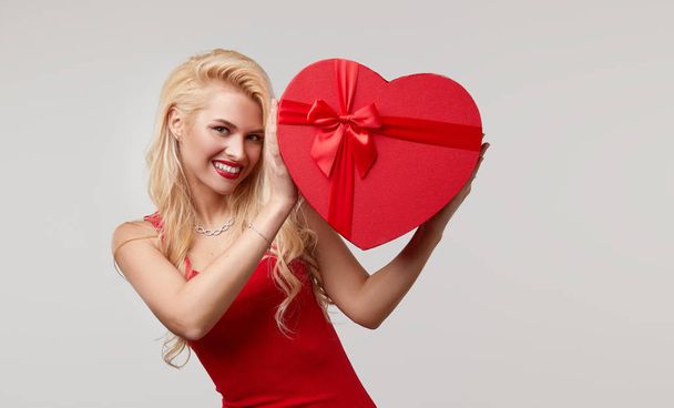 A young woman in a red dress holding a red heart shaped box near his face. Valentines Day and March 8 - Φωτογραφία, εικόνα