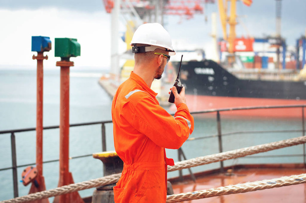 Um marinheiro experiente em macacão laranja e um capacete branco fala em um walkie-talkie e dá instruções para outros trabalhadores
. - Foto, Imagem