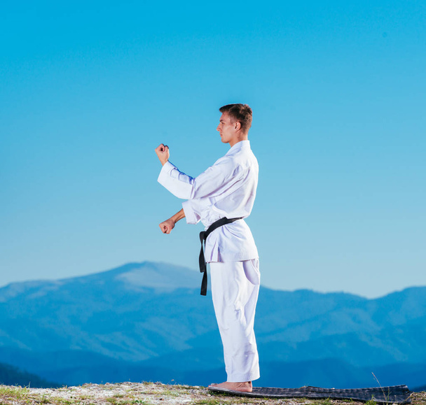 El atleta de karate rubio hace kata en la cima de una montaña mientras realiza una alineación de patadas, puñetazos y bloques en la cima de una montaña en un día soleado. - Foto, Imagen