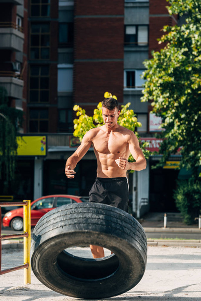 Joven deportista de mierda enfocado volteando neumáticos pesados en el lugar urbano - Foto, imagen
