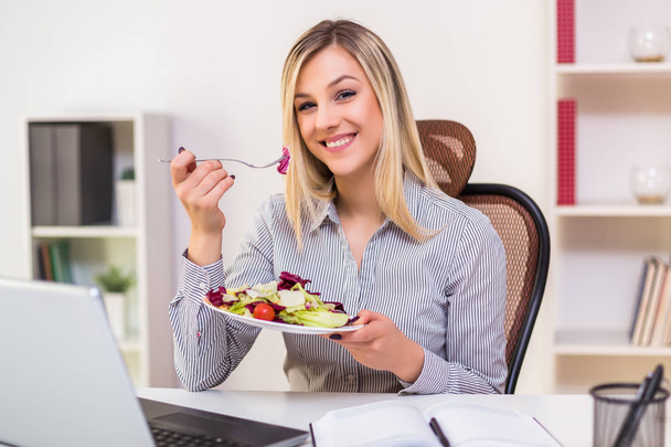 Beautiful businesswoman enjoys eating salad while working in her office. - Foto, imagen