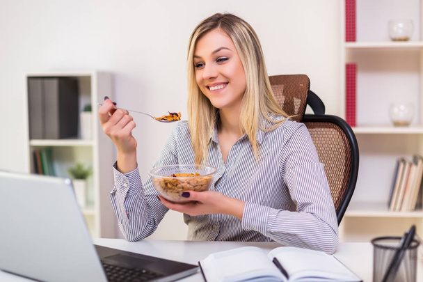 Beautiful businesswoman enjoys eating corn flakes for breakfast while working in her office. - Фото, изображение