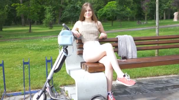 Feliz sorrindo jovem morena mulher sentar no banco perto de scooter elétrico e olhar para a câmera. Cena de aproximação
  - Filmagem, Vídeo
