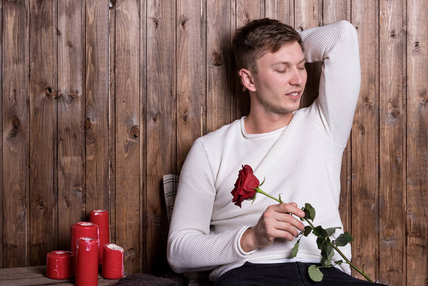 Young handsome man with a rose in his hands sitting near wood wall - Valokuva, kuva