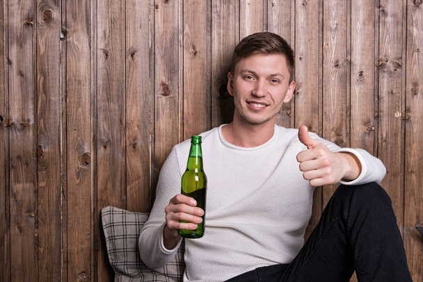 Young handsome man sitting near wood wall and drinking beer - Photo, image