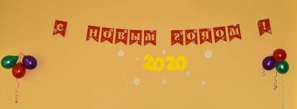 L'iscrizione in lettere rosse sulla parete arancione "Felice anno nuovo!". Lettere cirilliche ritagliate di carta rossa e gialla. 2020 scolpito sotto forma di formaggio giallo. Palloncini d'aria
. - Foto, immagini
