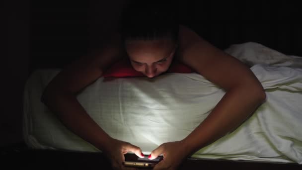 A young woman is lying on the bed and flipping through the various news in a mobile phone before bedtime. - Video