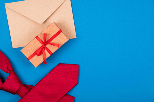 Composition of red neck tie, gift box with bow and brown craft paper envelope on blue cyan background. - Photo, image