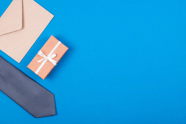 Composition of grey silver neck tie, gift box with white ribbon and brown craft paper envelope on blue cyan background. - Photo, Image