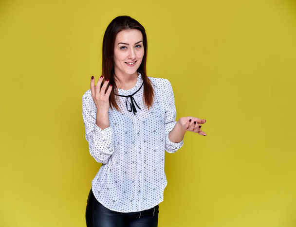 The concept of an office worker, teacher, manager. Portrait of a pretty brunette girl in a white business blouse smiling, talking to the camera on a yellow background. - 写真・画像
