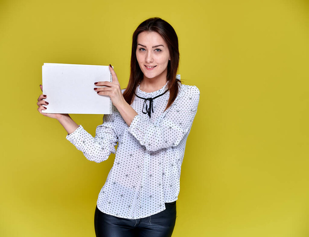 The concept of an office worker, teacher, manager. Portrait of a pretty brunette girl in a white business blouse smiling, talking to the camera on a yellow background with a folder in her hands. - Photo, image