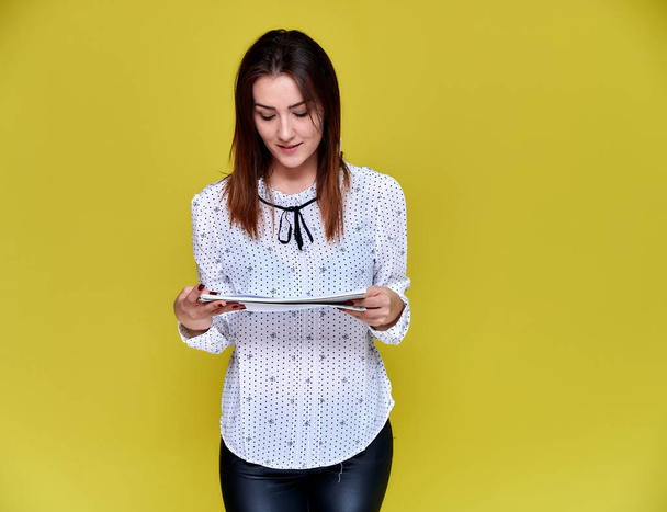 The concept of an office worker, teacher, manager. Portrait of a pretty brunette girl in a white business blouse smiling, talking to the camera on a yellow background with a folder in her hands. - Fotografie, Obrázek
