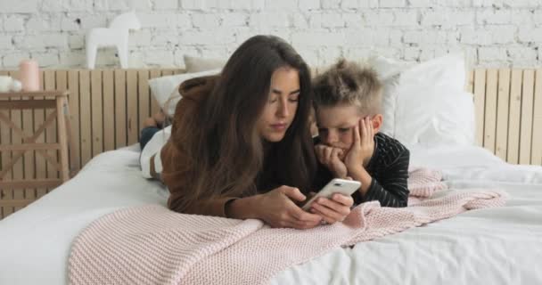 Mom and son browsing smartphone laying on bed in nursery, child is boring. - Πλάνα, βίντεο