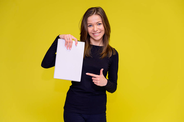 The concept of a smiling cute girl with a white folder in her hands. Portrait of a young woman in a black T-shirt stands in different poses with long hair on a yellow background. Shows hands, talking. - Fotografie, Obrázek