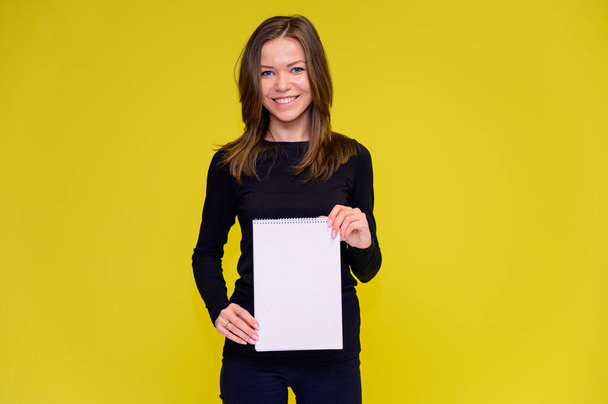 The concept of a smiling cute girl with a white folder in her hands. Portrait of a young woman in a black T-shirt stands in different poses with long hair on a yellow background. Shows hands, talking. - Valokuva, kuva