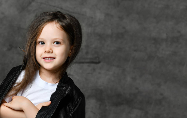 kleines lächelndes nettes Mädchen im schwarz-weißen Rockstar-Stil in lässiger Kleidung und weißen Turnschuhen vor grauem Betonhintergrund - Foto, Bild