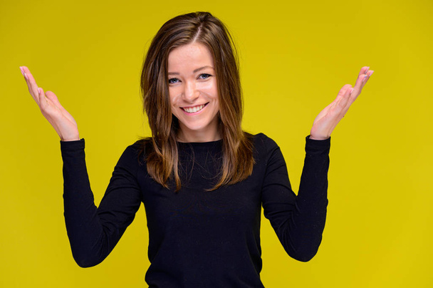 The concept of a smiling cute girl with positive emotions. Portrait of a young woman in a black T-shirt stands in different poses with long hair on a yellow background. Shows hands, talking. - Foto, Bild