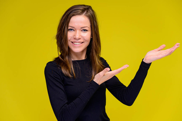 The concept of a smiling cute girl with positive emotions. Portrait of a young woman in a black T-shirt stands in different poses with long hair on a yellow background. Shows hands, talking. - Fotó, kép