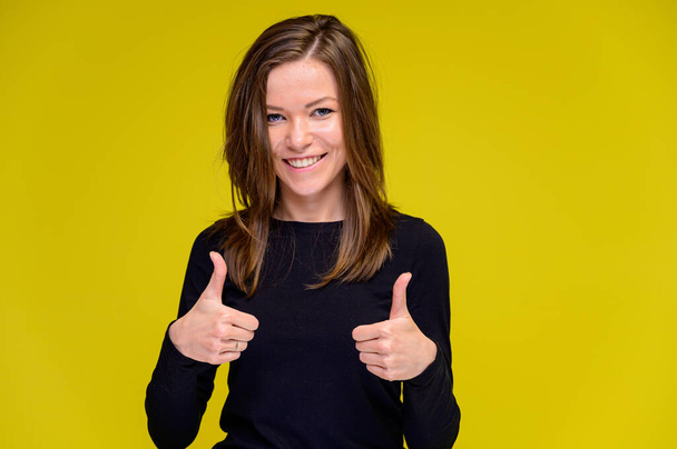 The concept of a smiling cute girl with positive emotions. Portrait of a young woman in a black T-shirt stands in different poses with long hair on a yellow background. Shows hands, talking. - Zdjęcie, obraz