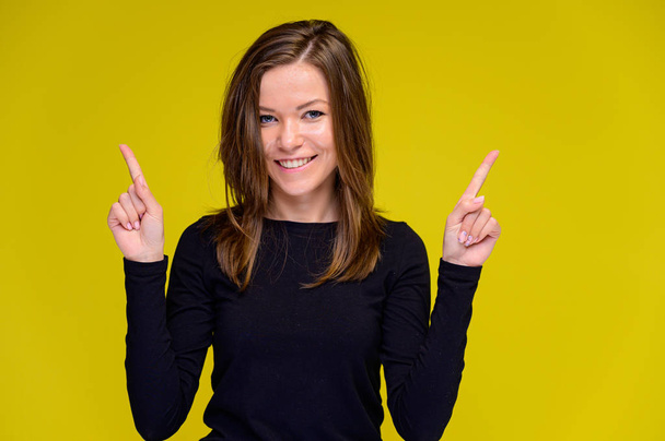 The concept of a smiling cute girl with positive emotions. Portrait of a young woman in a black T-shirt stands in different poses with long hair on a yellow background. Shows hands, talking. - Fotografie, Obrázek