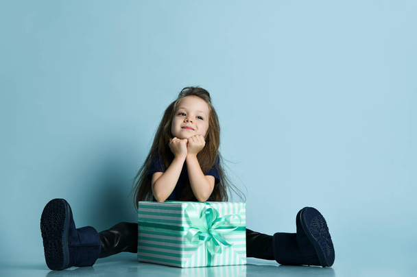 Small smiling girl in t-shirt, leather leggins and dark blue ugg boots sitting on floor with big present box - 写真・画像