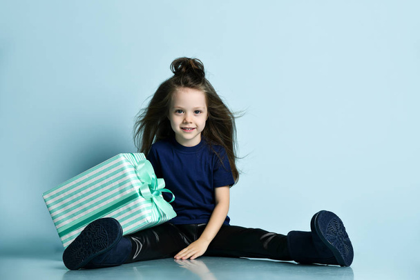 Small smiling girl in t-shirt, leather leggins and dark blue ugg boots sitting on floor with big present box - Fotoğraf, Görsel