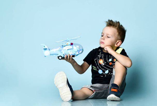 Szőke fiú elegáns alkalmi ruházat ül a padlón, és játszik játék helikopter jelen kék háttér - Fotó, kép