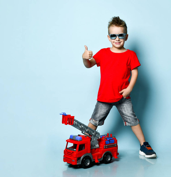 Pozitív szőke fiú fényes elegáns alkalmi ruházat, tornacipő, napszemüveg mellett álló játék tűzoltó motor és mutató hüvelykujj jel - Fotó, kép