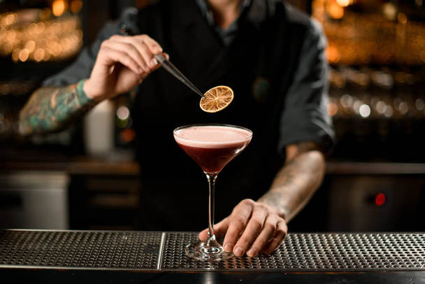 Professional male bartender decorating a delicious alcoholic cocktail in the glass with a dried orange with tweezers - Foto, Imagen