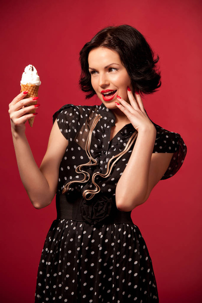 Young woman in black vintage dress standing eating ice cream over red background - Zdjęcie, obraz