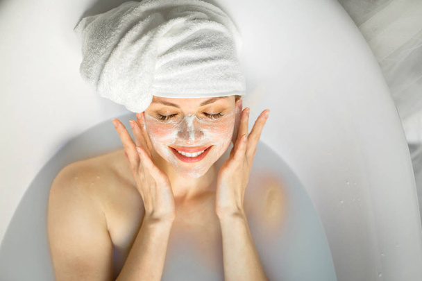 όμορφη νεαρή γυναίκα με μια πετσέτα στο κεφάλι της, βρίσκεται σε ένα μπάνιο με γάλα - Φωτογραφία, εικόνα