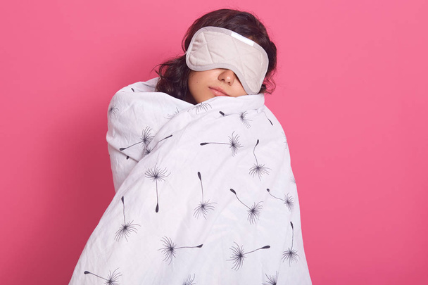 Sleep equipment concept. Portrait of brunette woman wrapped white blanket and wearing sleeping eye mask. Studio shot of young female isolated over pink background, lady being ready to fall asleep. - 写真・画像