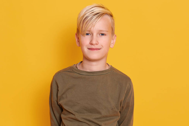 Close up portrait of liitle blond male child with charming smile wearing casual clothing standing against yellow studio wall, being photographed by his parents, looking at camera. Childhood concept. - Fotó, kép